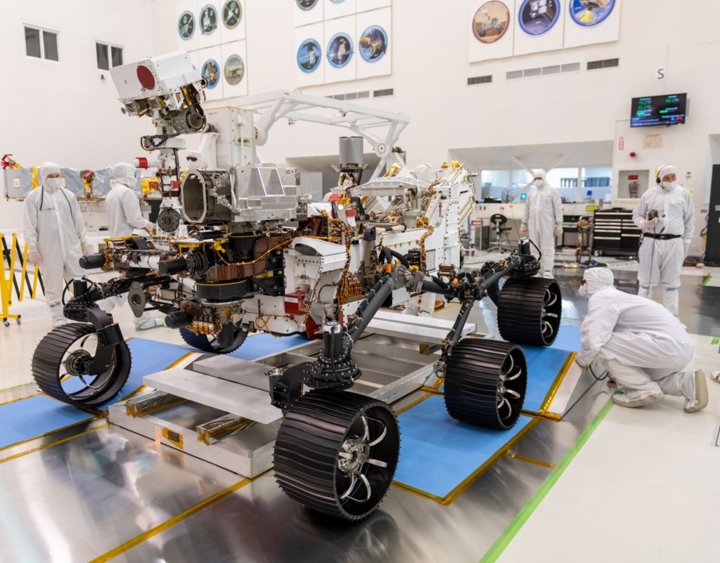 NASA's Perseverance six-wheeled rover pictured in a clean-room testing facility before launch, with engineers nearby.